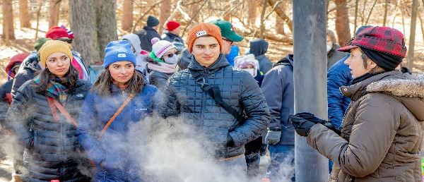 Volunteer speaking to visitors in front of maple syrup evaporator with steam rising.