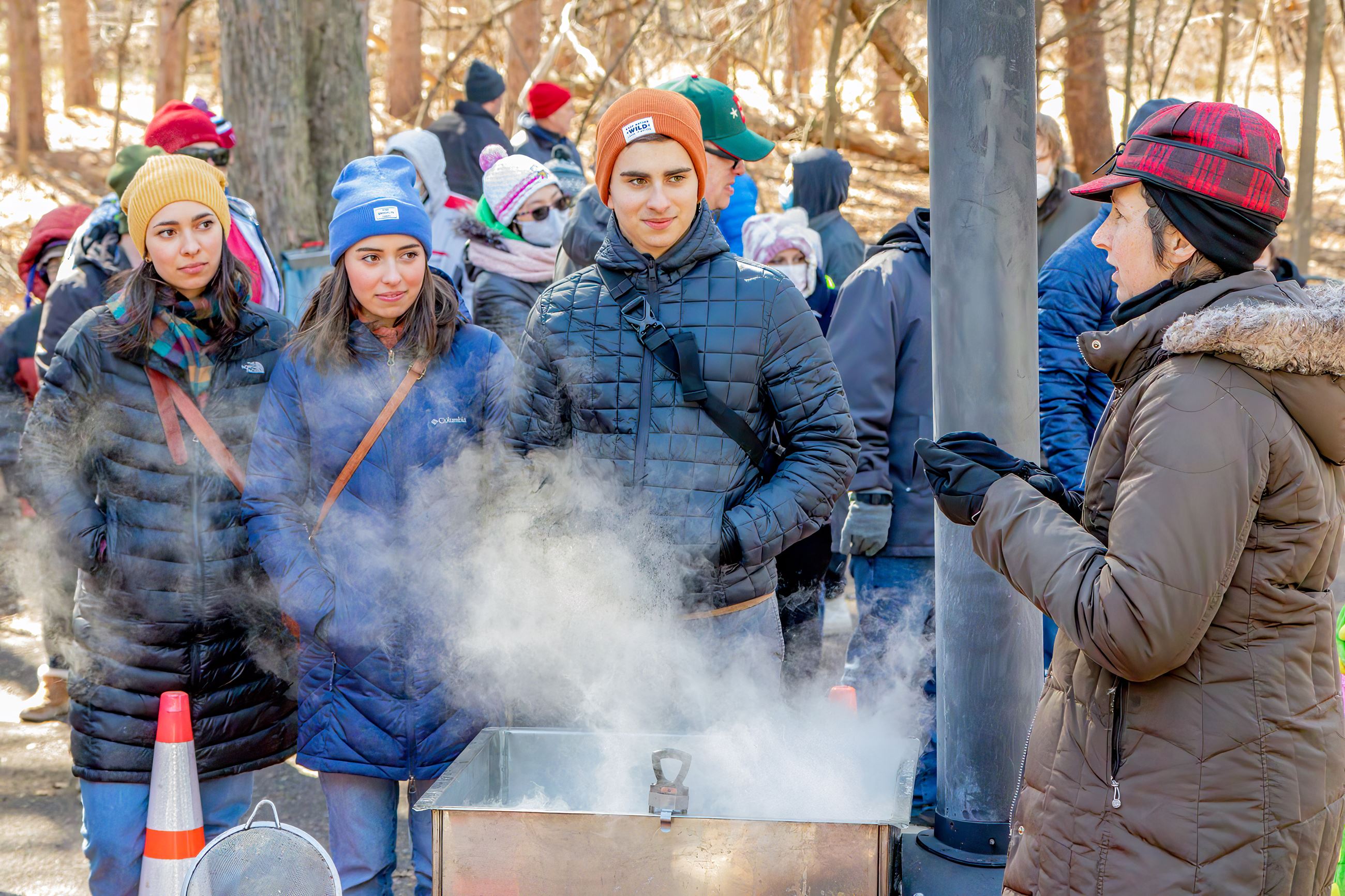 Volunteer speaking to visitors in front of maple syrup evaporator with steam rising.